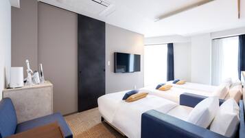 Una habitación de hotel con dos camas, un televisor montado en la pared, un escritorio con lámpara y una zona de estar con una silla y una mesita.
