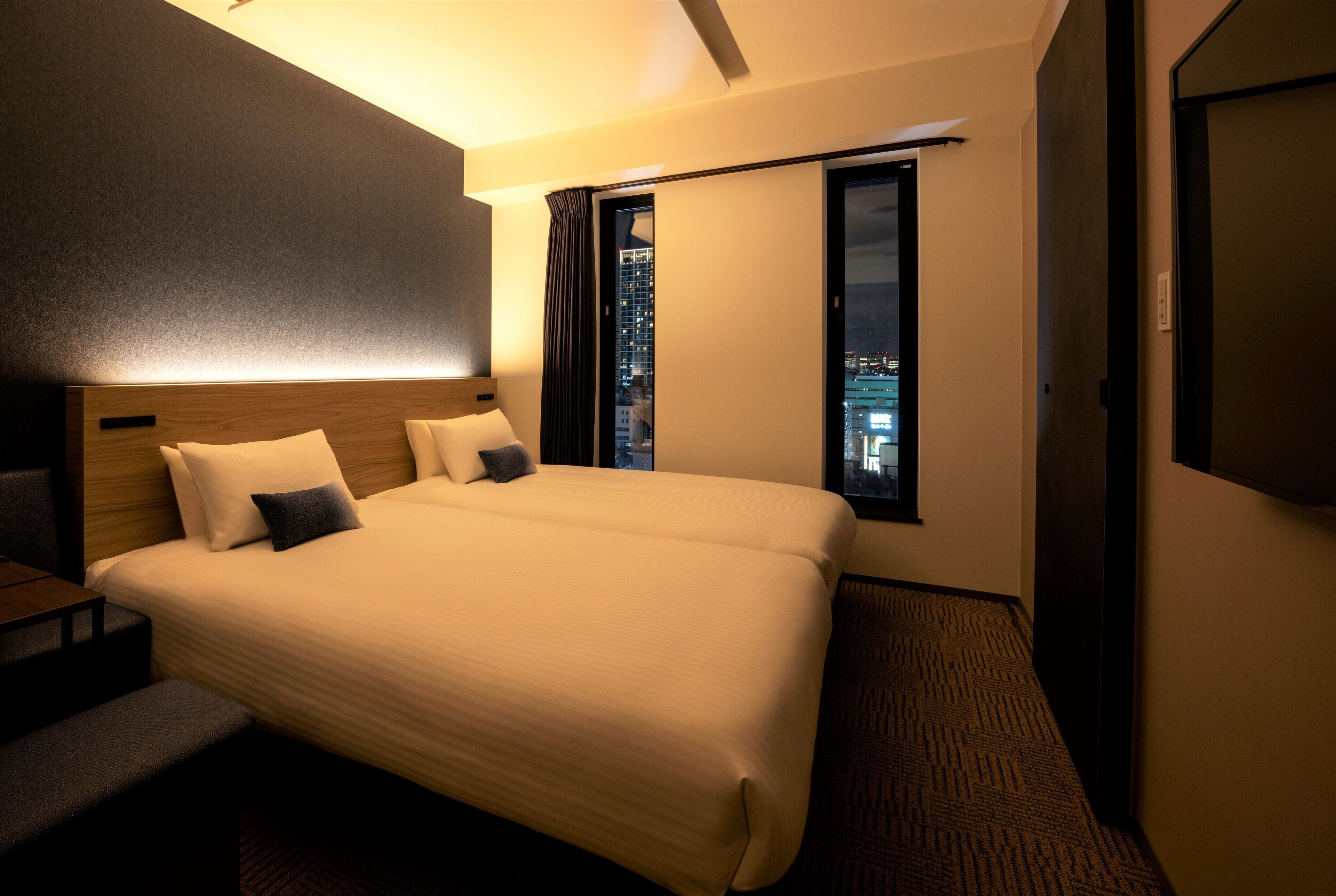 A hotel room with a large bed, a desk, and a view of a cityscape through a window.