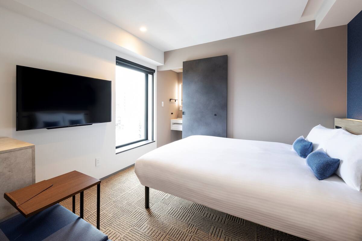 Une chambre d’hôtel moderne dotée d’un grand lit, d’un téléviseur à écran plat fixé au mur, d’une petite table en bois et d’une fenêtre laissant entrer la lumière naturelle.