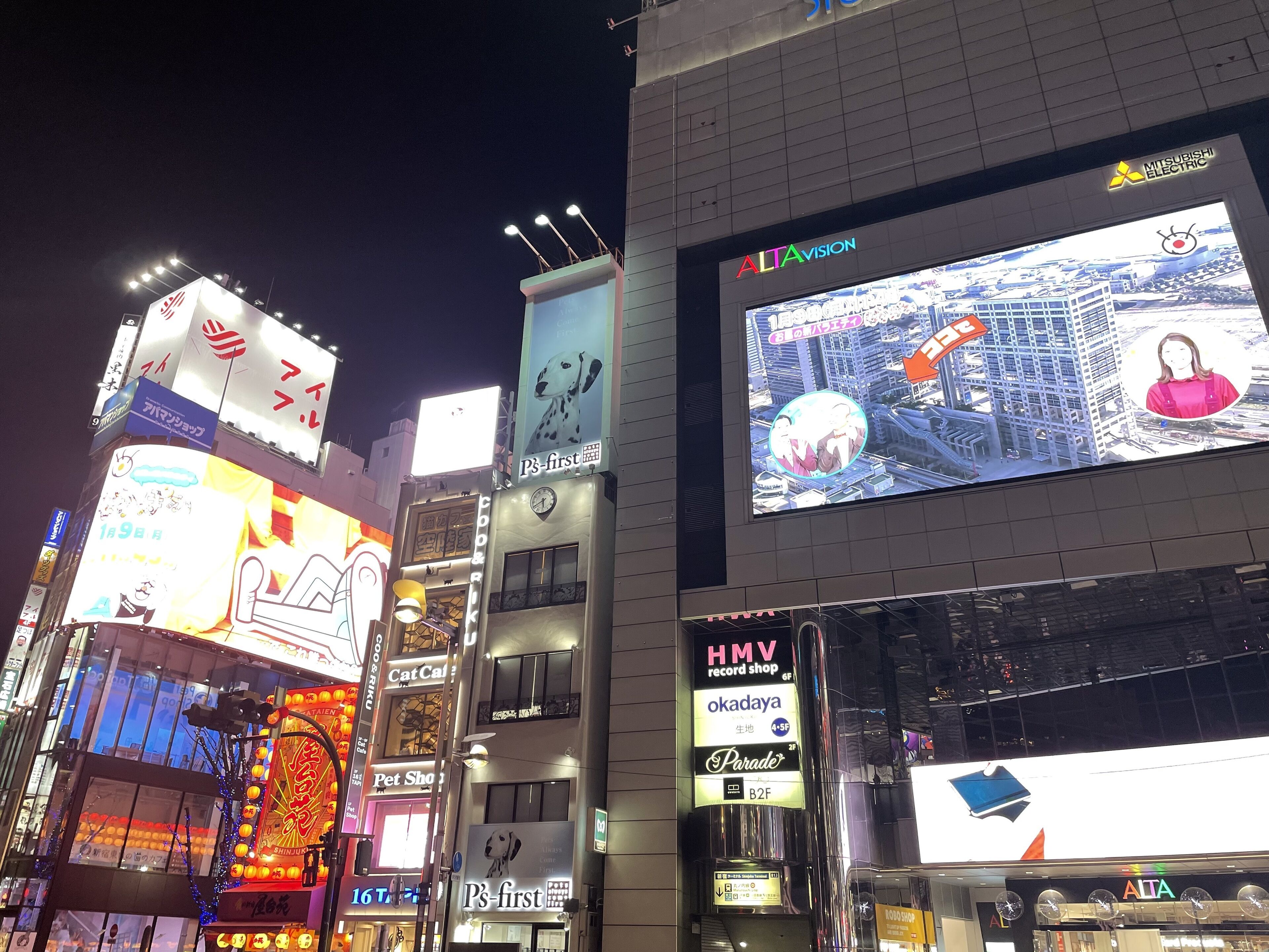 A brightly lit urban street at night with illuminated billboards and signs, including a large screen displaying a cityscape and advertisements.