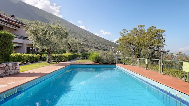 A swimming pool with a view of a mountain, surrounded by greenery and a building. - Apartment 'Assenza 9' with Lake View, Wi-Fi and Air Conditioning (Brenzone sul Garda)