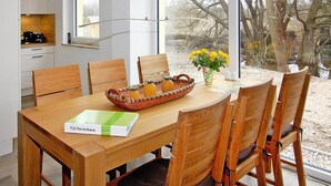 Ein Essbereich mit einem Holztisch, sechs Stühlen, einem Korb voller Ananas, einer Blumenvase und einem Fenster mit Blick auf Bäume und ein Gebäude.