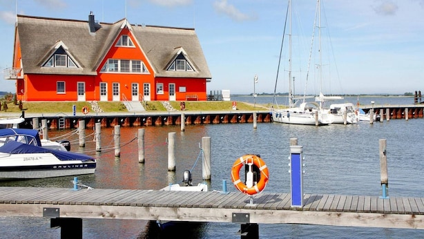 Ein rotes Gebäude mit einem Steg, der in ruhiges Wasser führt, einem Rettungsring und einem Segelboot.