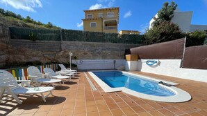 Piscina com deck revestido, cadeiras de descanso brancas e parede de pedra.