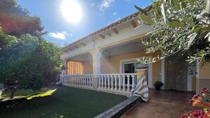 Uma casa com um pátio ladrilhado, um pequeno jardim com grama sintética e uma varanda com corrimão branco.