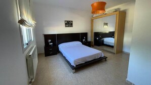Um quarto com cama, mesas de cabeceira, radiador, janela com cortinas e um espelho.
