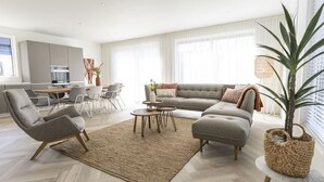 Een moderne woonkamer met een hoekbank, een grijze fauteuil, een salontafel en een eethoek met stoelen en een tafel.