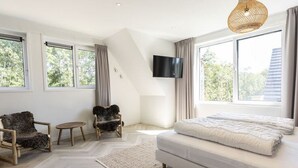 Een moderne slaapkamer met een groot bed, een aan de muur bevestigde televisie en uitzicht op het groen door grote ramen.