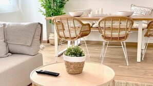 Un salon moderne avec une table basse ronde en bois, des fauteuils en osier, un canapé et une table à manger avec des chaises. Deux lampes en osier sont suspendues au-dessus de la table à manger, et une plante en pot est posée sur la table basse.
