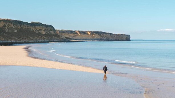 Ein Strand mit einer Person, die am Ufer entlanggeht, Klippen im Hintergrund und ruhigem Wasser.