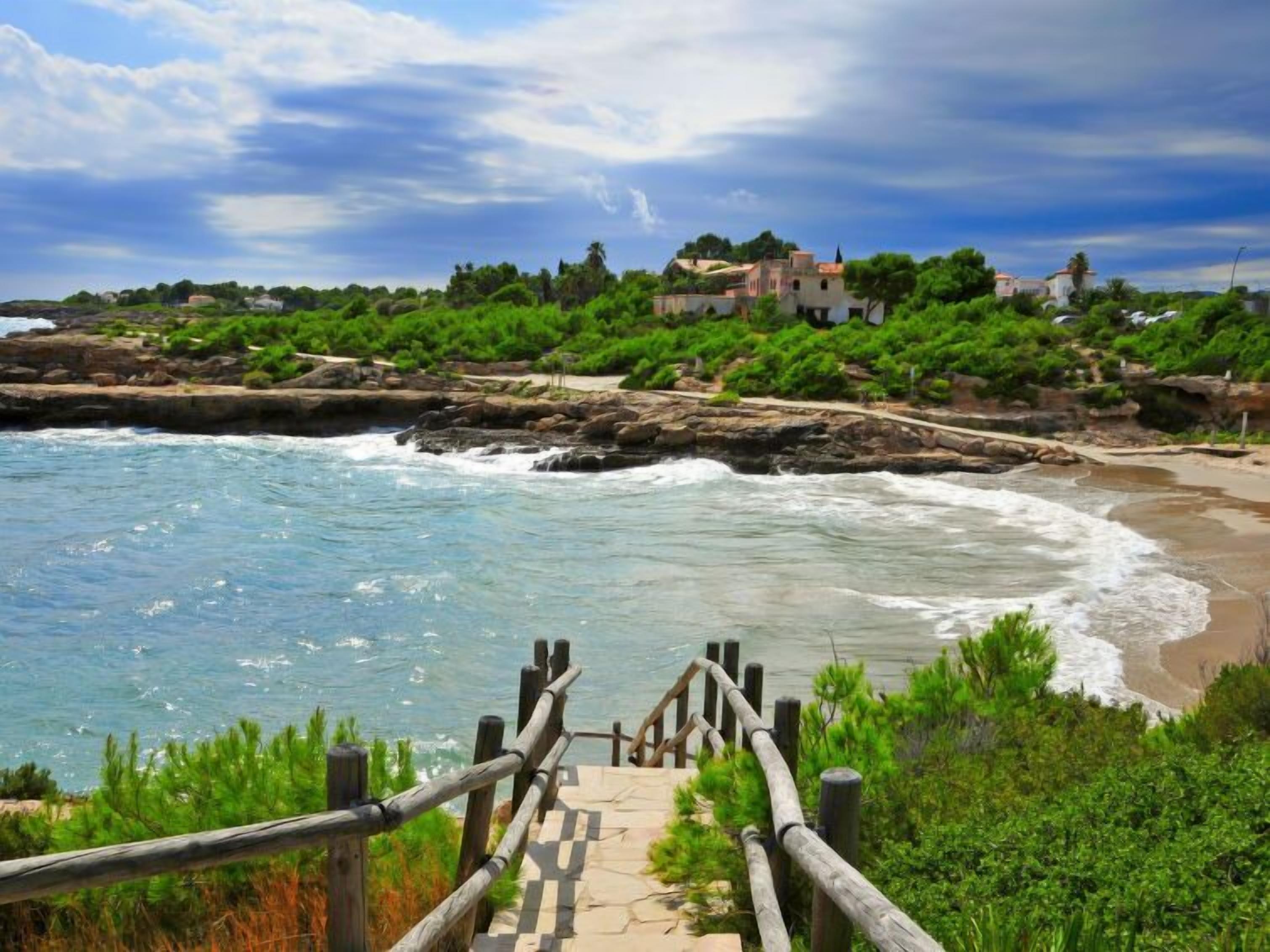 A coastal walkway leading to a beach with rocky outcrops and a view of a town in the distance.