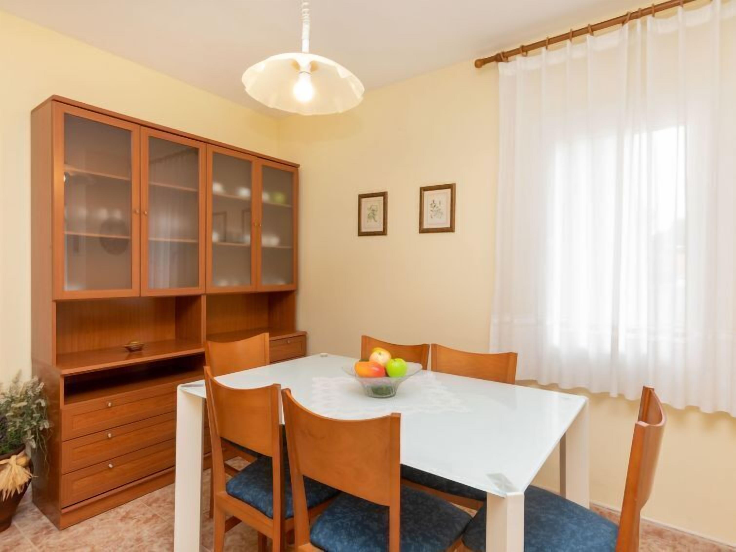 A dining area with a white table, wooden chairs, a cabinet with glass doors, and a ceiling light.
