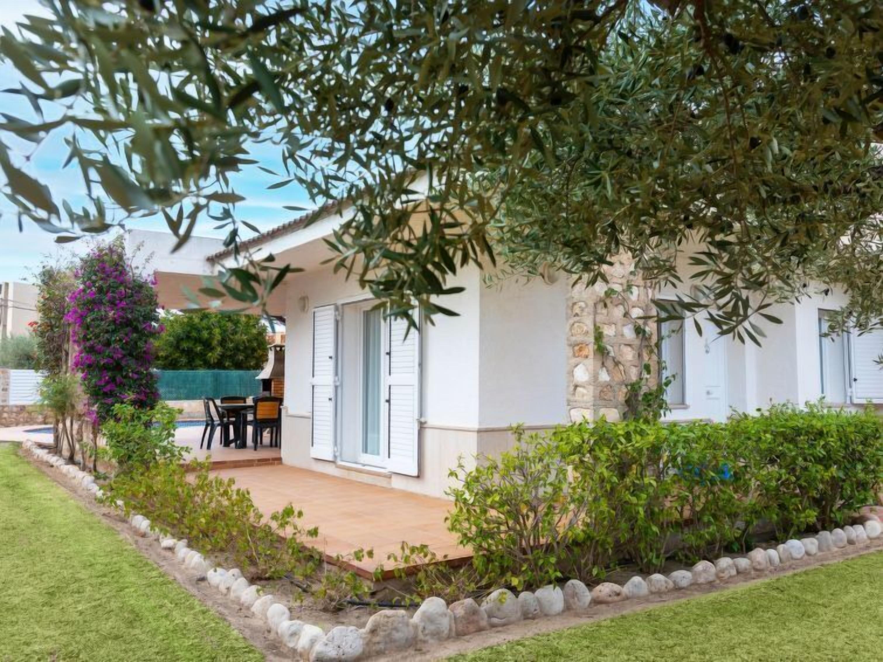 A white house with a stone facade, a patio with outdoor furniture, and a garden with various plants and a purple flowering bush.