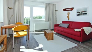 A modern living room with a red sofa, yellow chairs, a wooden coffee table, and a view of the outdoors through large windows. - Ferienwohnung, St. Peter-Ording (St. Peter-Ording)
