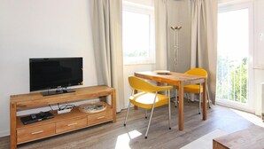 A modern room with a TV on a wooden stand, a dining table with yellow chairs, and large windows with curtains. - Ferienwohnung, St. Peter-Ording (St. Peter-Ording)