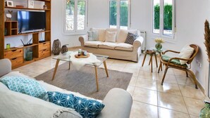 Una sala de estar con sofá, una mesa de centro, un televisor sobre un soporte de madera y un ventanal con vistas a zonas verdes.