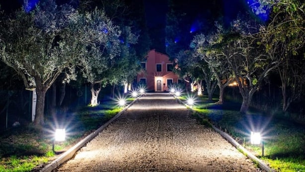 Un sentiero conduce a una casa con facciata rossa, circondata da alberi e illuminata da lampioni.