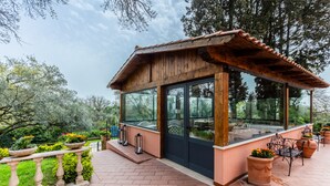 Un gazebo in legno con ampie finestre e tetto in tegole, circondato da piante in vaso e un patio in mattoni.