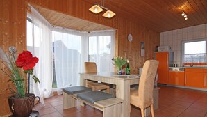 A wooden interior with a dining table, benches, a chair, a kitchen area, and a window with curtains.