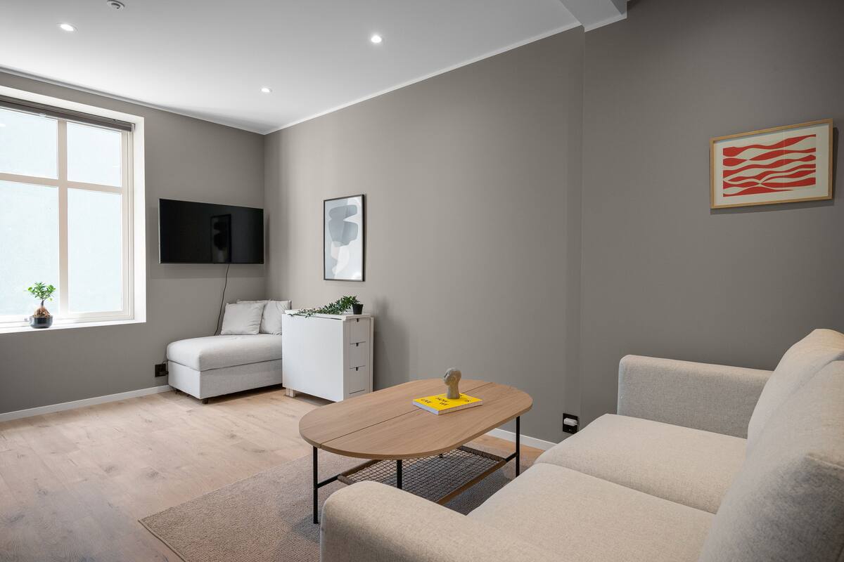 Een moderne woonkamer met een bank, een salontafel, een aan de muur bevestigde televisie en een raam met een plant op de vensterbank.