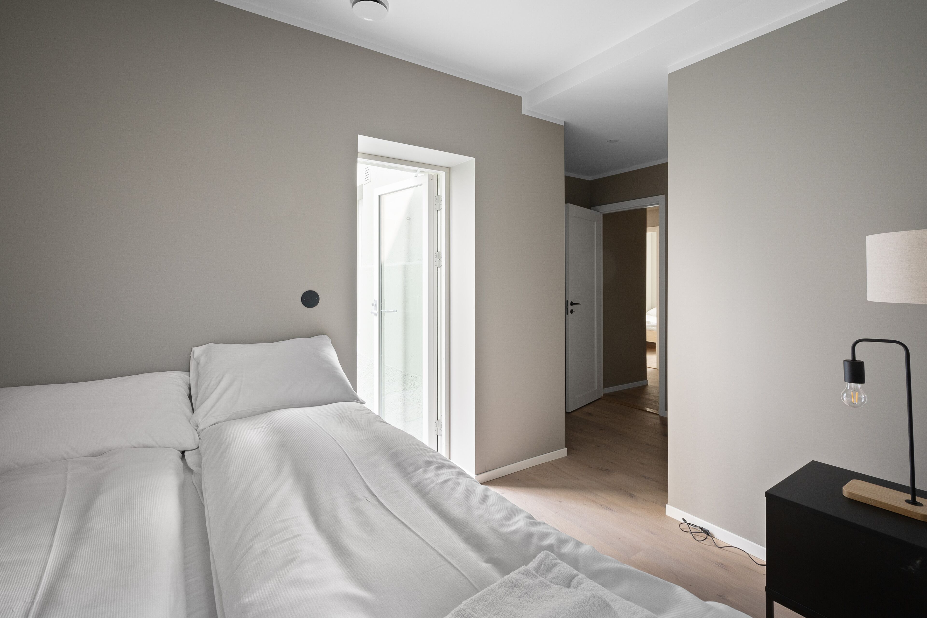 A bedroom with a bed, a bedside table with a lamp, and a door leading to another room.