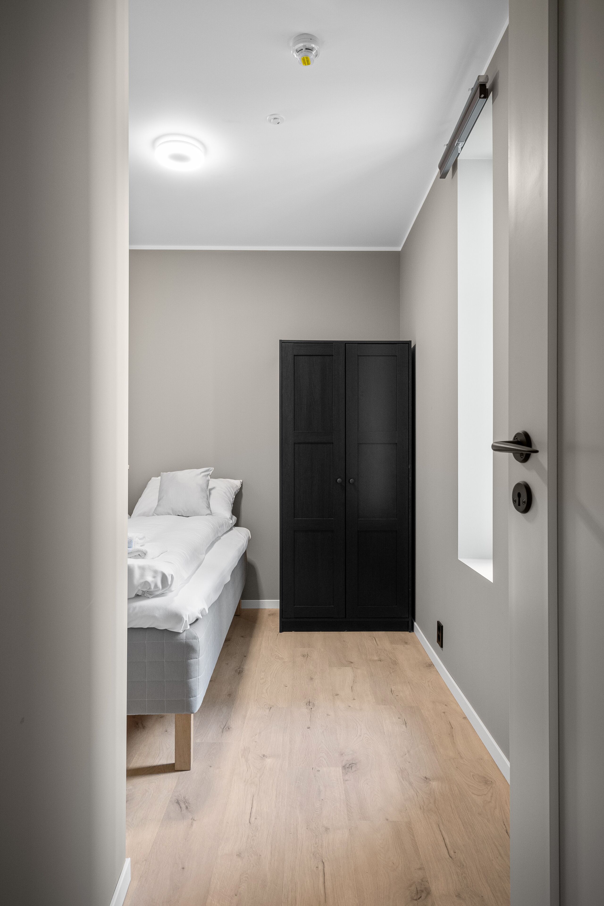 A narrow room with a bed, a dark wardrobe, and a door with a handle.