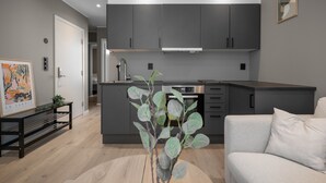 Eine moderne Küche mit dunklen Schränken, einer Edelstahl-Dunstabzugshaube und einem eingebauten Backofen. Neben der Tür befindet sich eine Bank mit einer Pflanze, und im Vordergrund ein helles Sofa.