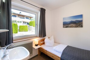 Ein kleiner, ordentlicher Raum mit einem Bett, einem Fenster mit Vorhängen, einem Heizkörper, einem Waschbecken und einem Bild an der Wand.