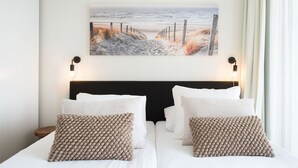 A hotel room with two beds, a headboard, wall-mounted lamps, and a beach scene painting. - Apartment in Netherlands near the Beach (Den Haag)