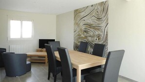A modern dining area with a wooden table and black chairs, a large abstract wall art, a window with blinds, and a white radiator. - House in Saint-Quay near 7 Beaches (St. Quay-Portrieux)