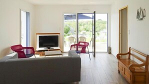 Una sala de estar moderna con un sofá gris, un banco de madera, un televisor y un gran ventanal corredizo que da a un espacio exterior con sillas.