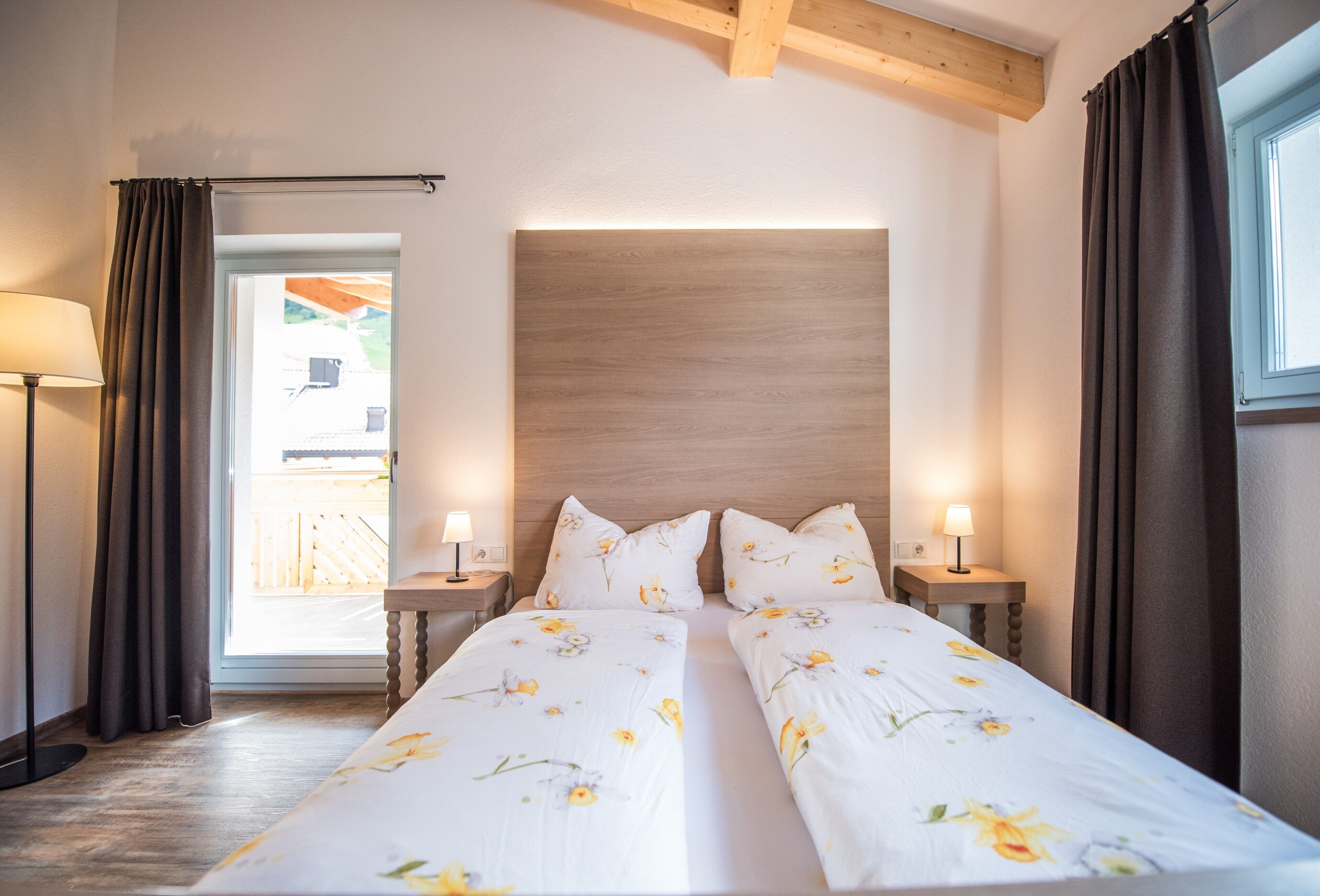 Ein Doppelbett mit floraler Bettwäsche, zwei Nachttischlampen, ein Holzkopfstück und ein Blick durch eine Tür in einen Außenbereich.