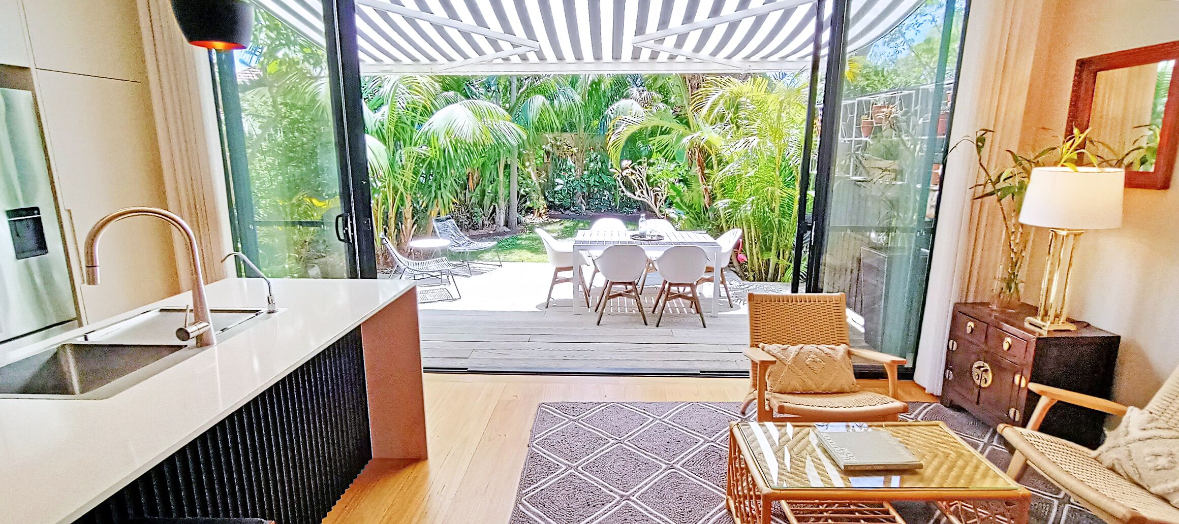 A modern kitchen with a sink, a dining area with chairs and a table, and a view of a lush garden through glass doors.