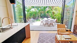 A modern kitchen with a sink, a dining area with chairs and a table, and a view of a lush garden through glass doors.
