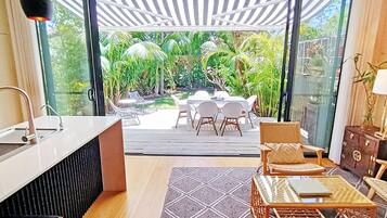A modern kitchen with a sink, a dining area with chairs and a table, and a view of a lush garden through glass doors.
