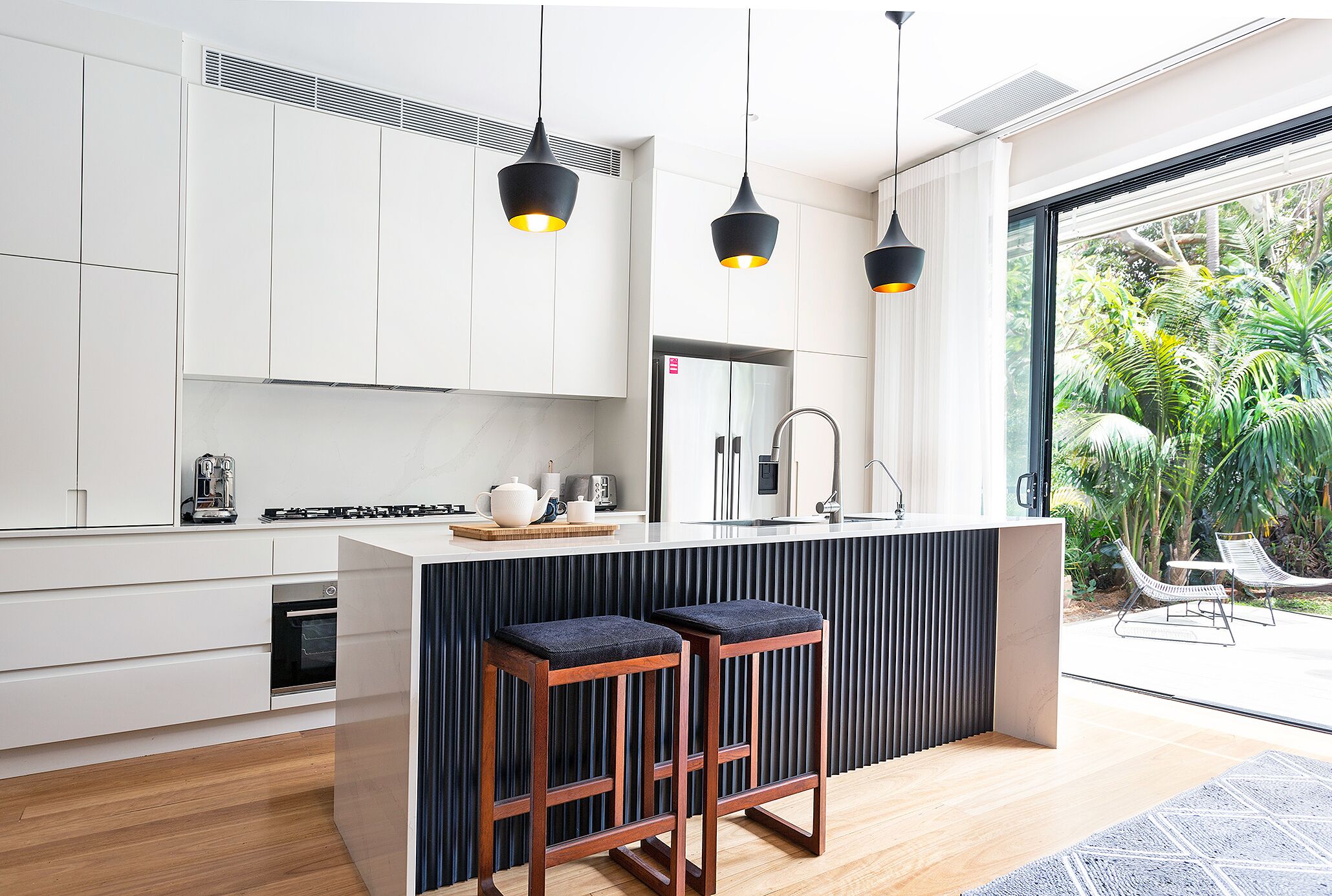 A modern kitchen with white cabinets, a central island with stools, pendant lighting, and a view of a lush garden.