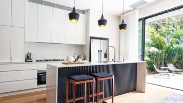 A modern kitchen with white cabinets, a central island with stools, pendant lighting, and a view of a lush garden.