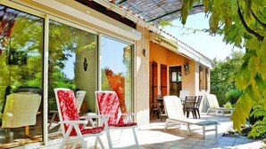 Un espacio de patio al aire libre con sillas y una mesa blancos, rodeado de puertas y ventanas de vidrio, y una zona de estar cubierta.