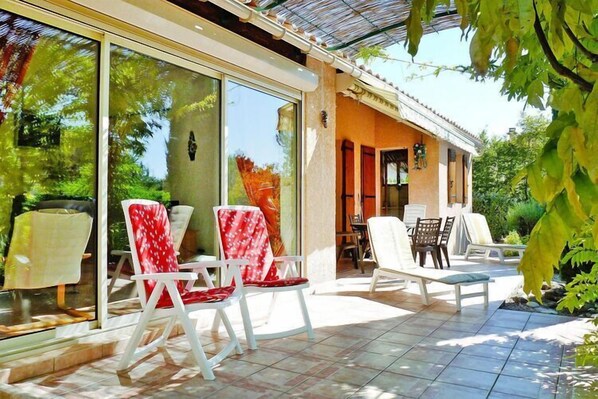 Un espacio de patio al aire libre con sillas y una mesa blancos, rodeado de puertas y ventanas de vidrio, y una zona de estar cubierta.