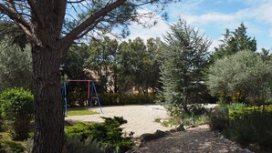 Un jardín con un camino de grava, un columpio y diversas árboles y arbustos.