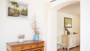 Una habitación con un aparador de madera, un florero con plantas secas, un cuadro de un paisaje y un mueble con un florero y una silla.