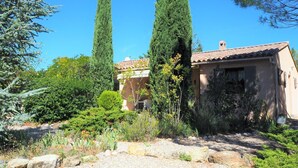 Un edificio de estilo mediterráneo con techo de tejas, rodeado de un jardín cuidado con cipreses y diversos arbustos.