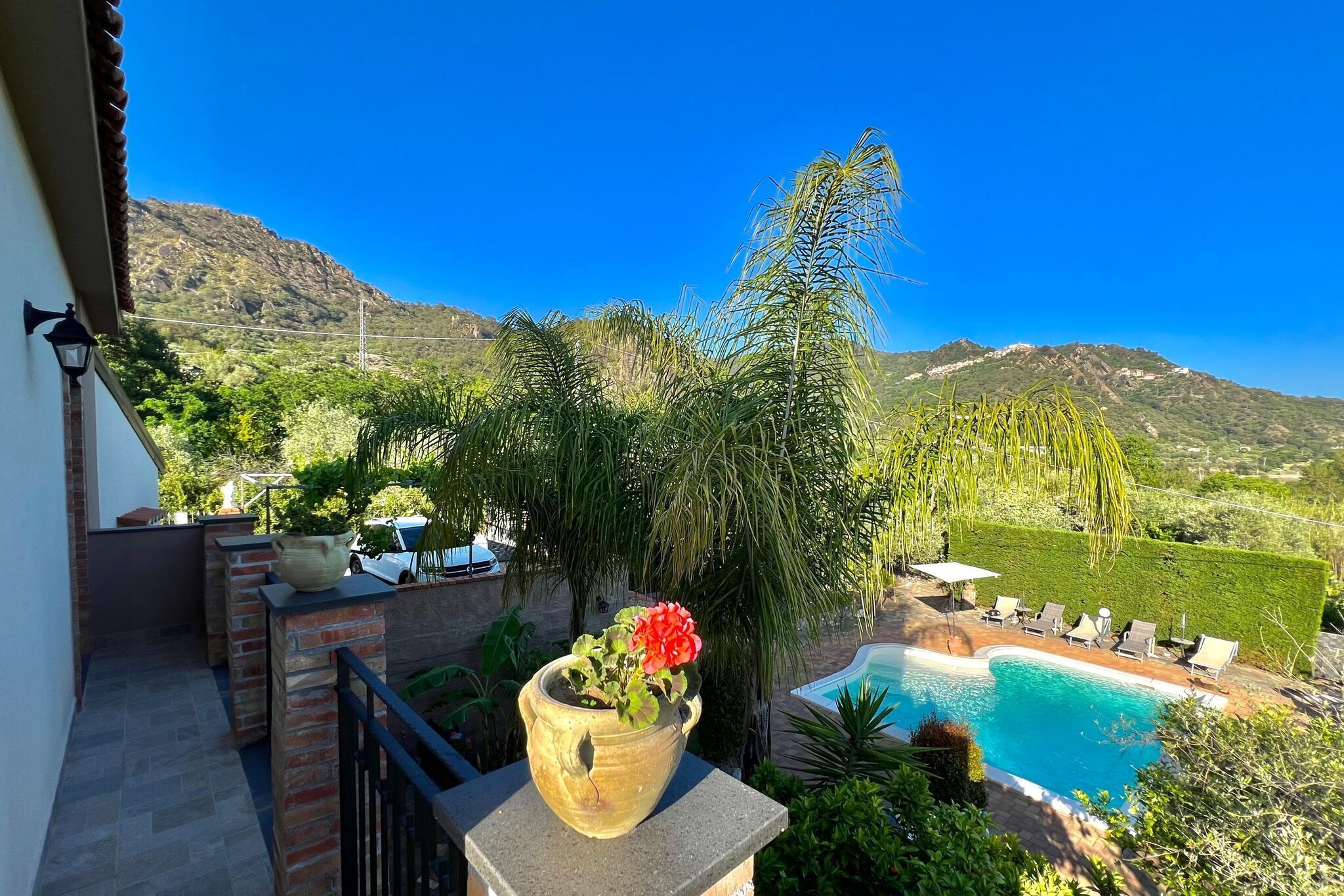 A poolside view with a mountain backdrop, a potted plant with red flowers, and a clear blue sky.