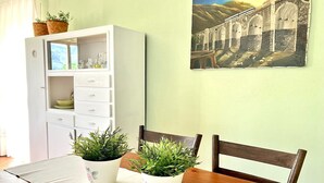 Een eetgedeelte met een tafel gedekt voor twee personen, twee potplanten, een witte kast met lades en een aan de muur hangend schilderij van een brug.
