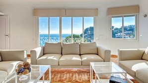 Ein Wohnzimmer mit einer Couch, zwei Sesseln, einem Glastisch und Meerblick durch große Fenster.