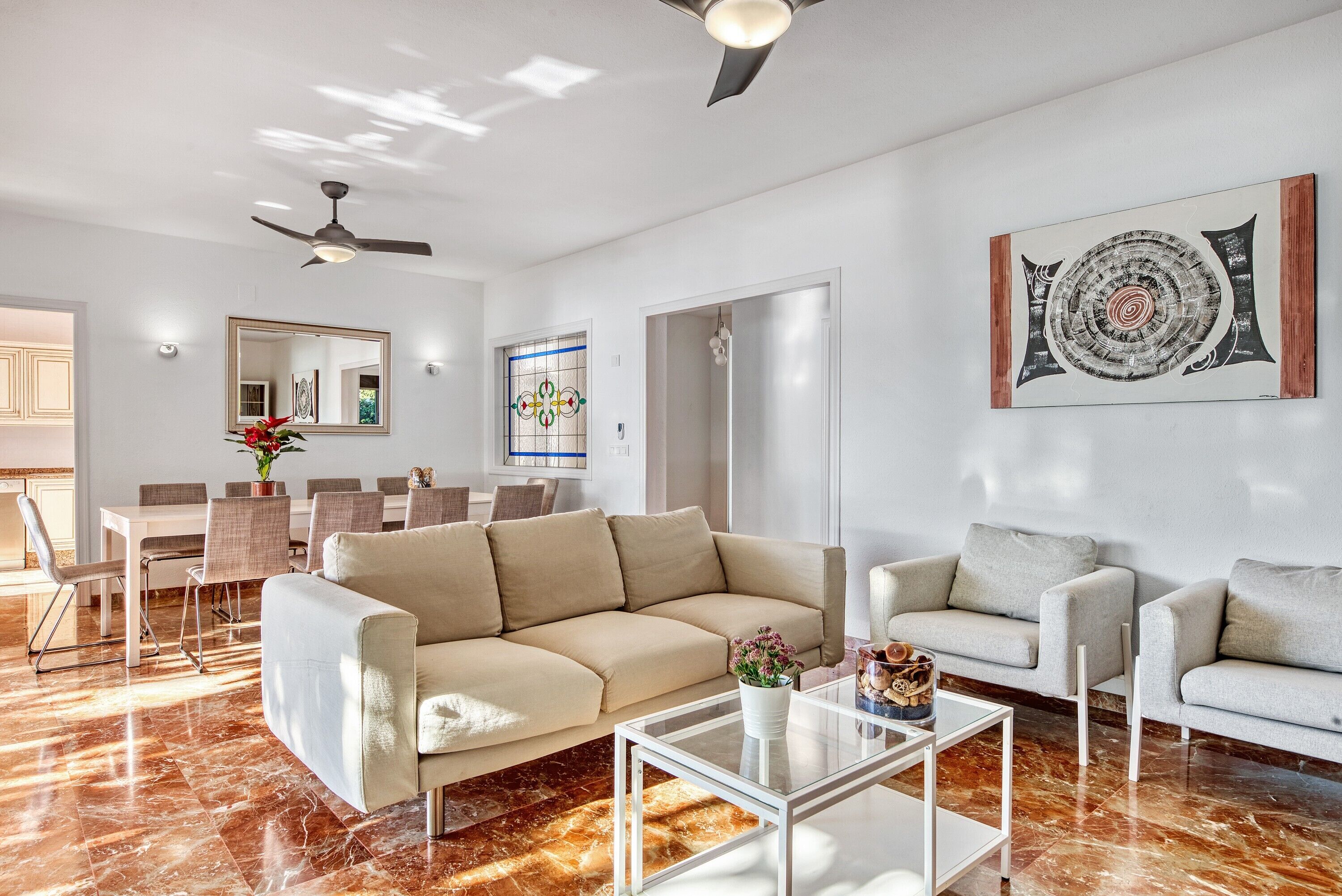 A modern living room with a beige sofa, a glass coffee table, and a dining area with a white table and chairs.