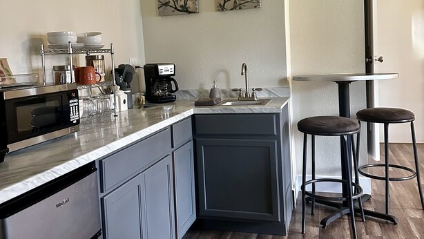 A compact kitchen with a built-in dishwasher, a coffee maker, and a sink. There are two stools and a small table.