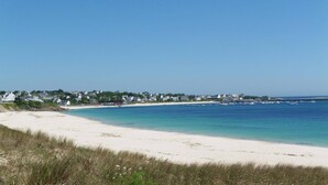 A coastal beach with white sand and clear blue water. There are houses along the shoreline.