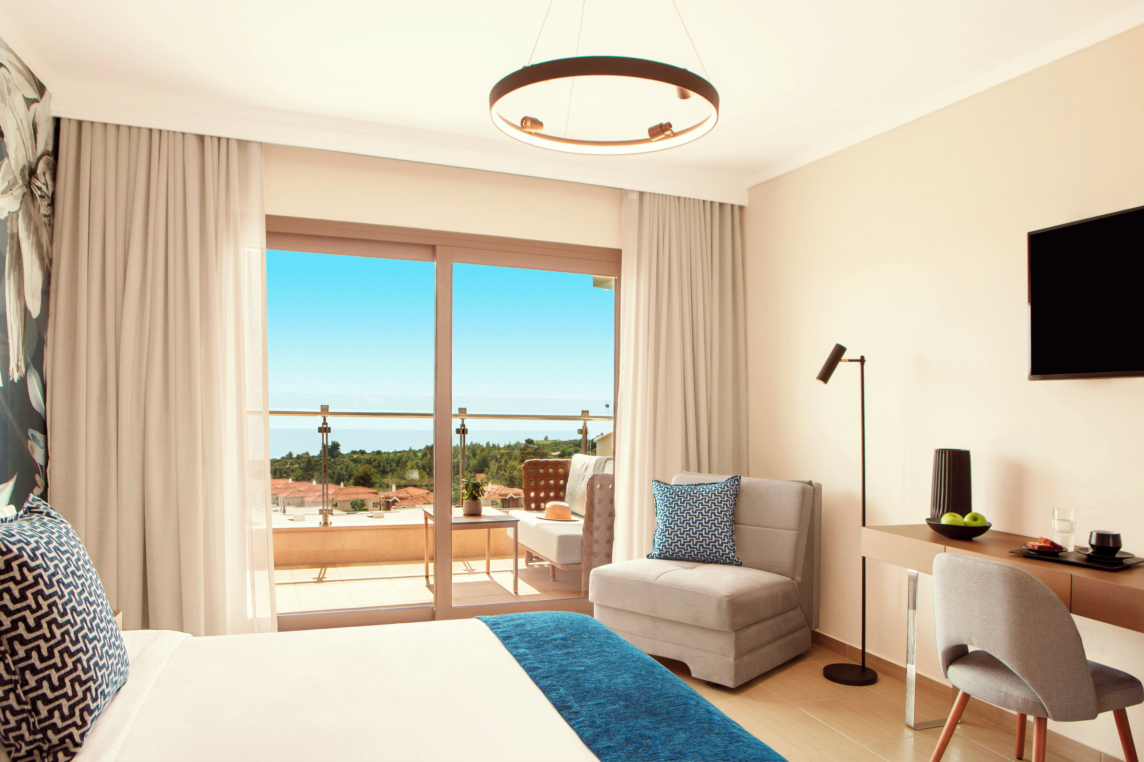 Quarto de hotel com uma cama grande, área de estar com sofá e poltrona, escrivaninha com cadeira, televisão na parede e varanda com vista para a cidade.