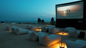 Cine al aire libre con una pantalla grande, asientos tipo sillón de mimbre y faroles.